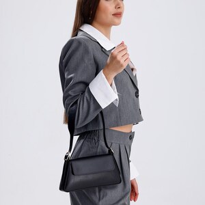 Puede incluir: Una mujer lleva un traje gris con una camisa blanca de cuello debajo. Lleva un bolso de mano de cuero negro con una correa de cadena dorada.