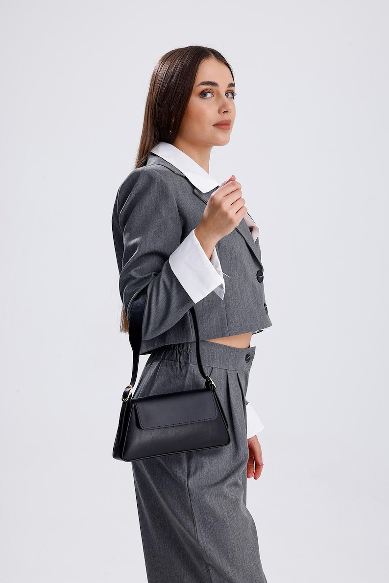 Puede incluir: Una mujer lleva un traje gris con una camisa blanca de cuello debajo. Lleva un bolso de mano de cuero negro con una correa de cadena dorada.