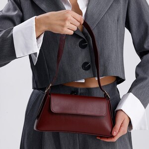 May include: A burgundy leather shoulder bag with a flap closure and a short strap. The bag is being held by a person wearing a grey blazer and white shirt.