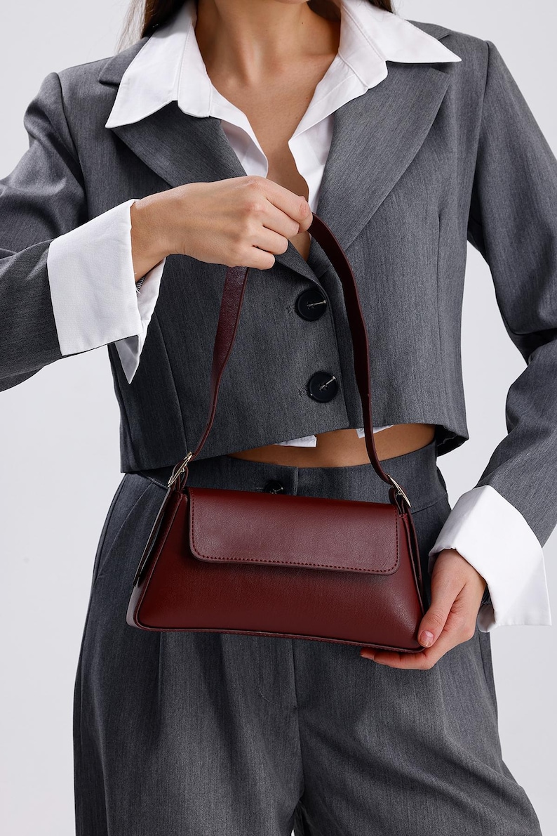 May include: A burgundy leather shoulder bag with a flap closure and a short strap. The bag is being held by a person wearing a grey blazer and white shirt.