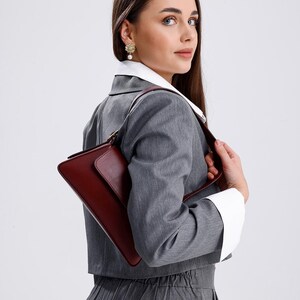 May include: A woman wearing a grey blazer and trousers with a white collared shirt underneath. She is holding a burgundy leather handbag with a gold clasp.