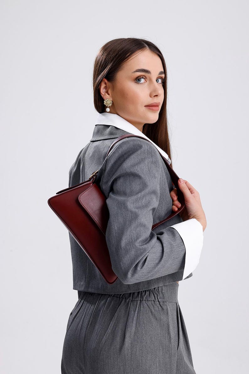 May include: A woman wearing a grey blazer and trousers with a white collared shirt underneath. She is holding a burgundy leather handbag with a gold clasp.