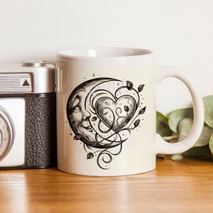 Könnte beinhalten: Weiße Keramiktasse mit einer schwarz-weißen Illustration eines zunehmenden Mondes mit einem Gesicht und einem Herzen, das mit Ranken verschlungen ist.