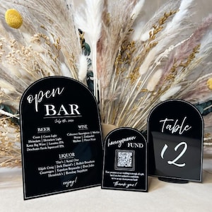 May include: Three black acrylic signs with white text. The first sign says "open bar" with a list of beer, wine, and liquor options. The second sign says "honeymoon fund" with a QR code. The third sign says "table 12".