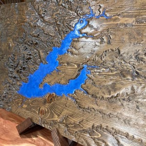 Könnte beinhalten: Ein Wandkunststück aus Holz mit einer blauen Harzeinlage, die einen See oder Fluss darstellt. Das Holz hat eine natürliche, rustikale Oberfläche.