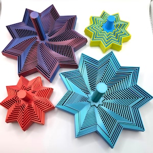May include: Four star-shaped objects in various colours, each with a central peg. The objects have a layered, geometric design. Colours include red, blue, green, and a purple-red gradient. These are likely decorative or functional items.