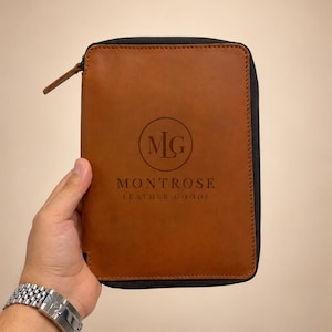 May include: A rectangular tan leather folio with a black zipper closure. The front features a circular logo with the letters MG inside, followed by the text MONTROSE LEATHER GOODS. The case has a smooth finish and a matching tan leather zipper pull.