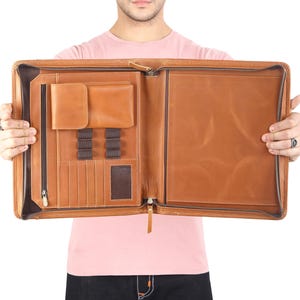 May include: A tan leather zippered portfolio case featuring an interior organizer with a zippered pocket, a flap-covered compartment, multiple card slots, and elastic pen loops on the left side, with a large open document sleeve on the right side.