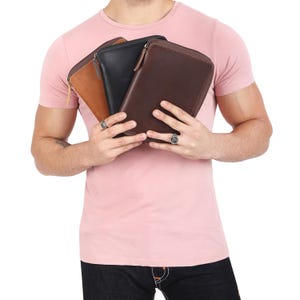 May include: Three leather wallets in brown, black, and tan with zippers. The wallets are held in the hands of a person wearing a pink t-shirt.