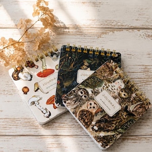 May include: Three square spiral-bound journals with gold wire binding. One journal features a white cover with mushroom illustrations, one has a dark forest pattern, and one displays owls. Each cover includes a white label with the text JOURNAL.