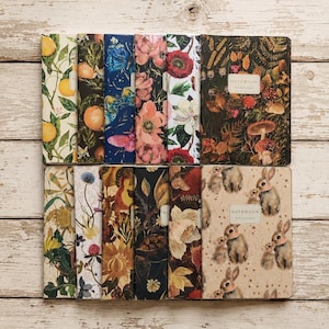 May include: A collection of twelve rectangular notebooks arranged in two rows, featuring diverse botanical, floral, and animal illustrations. Each cover displays unique patterns including oranges, butterflies, mushrooms, and rabbits, with some labeled NOTEBOOK.
