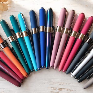 May include: A collection of fountain pens arranged in a fan shape, featuring a variety of matte and metallic finishes in colors including orange, teal, blue, pink, magenta, gray, white, and black, each with a gold-toned metal center band.
