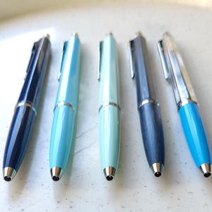 May include: Five pens in various shades of blue and teal are displayed. Each pen features a silver clip and tip. The colours range from dark navy to light turquoise, with a wood-grain textured pen in the middle. The pens are approximately 14 cm long.