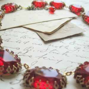 May include: A red glass necklace with gold accents. The necklace is made of faceted red glass beads with gold metal frames. The necklace is lying on a piece of paper with handwritten text.