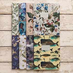 May include: A collection of eight notebooks featuring diverse artistic covers, including botanical prints with flowers and berries, deep sea themes with whales and seahorses, and mythical forest scenes with deer and unicorns, all labeled with the text NOTEBOOK.