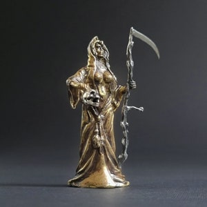 May include: A gold-colored figurine of a woman in a hooded robe, holding a skull in one hand and a scythe in the other. The figurine is standing on a black surface.