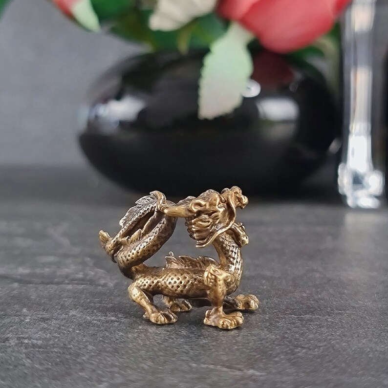 May include: A small, gold-colored metal dragon figurine with intricate details. The dragon is curled up with its tail in its mouth.