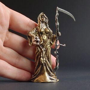 May include: A gold-colored metal figurine of a female figure in a hooded robe, holding a skull in one hand and a scythe in the other. The figurine is standing on a black surface.
