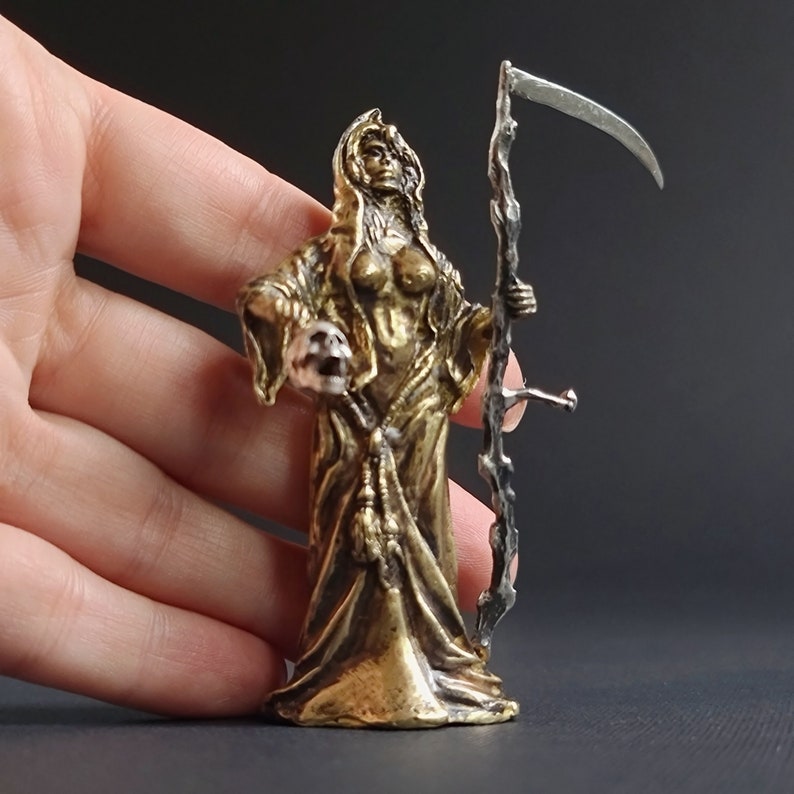 May include: A gold-colored metal figurine of a female figure in a hooded robe, holding a skull in one hand and a scythe in the other. The figurine is standing on a black surface.