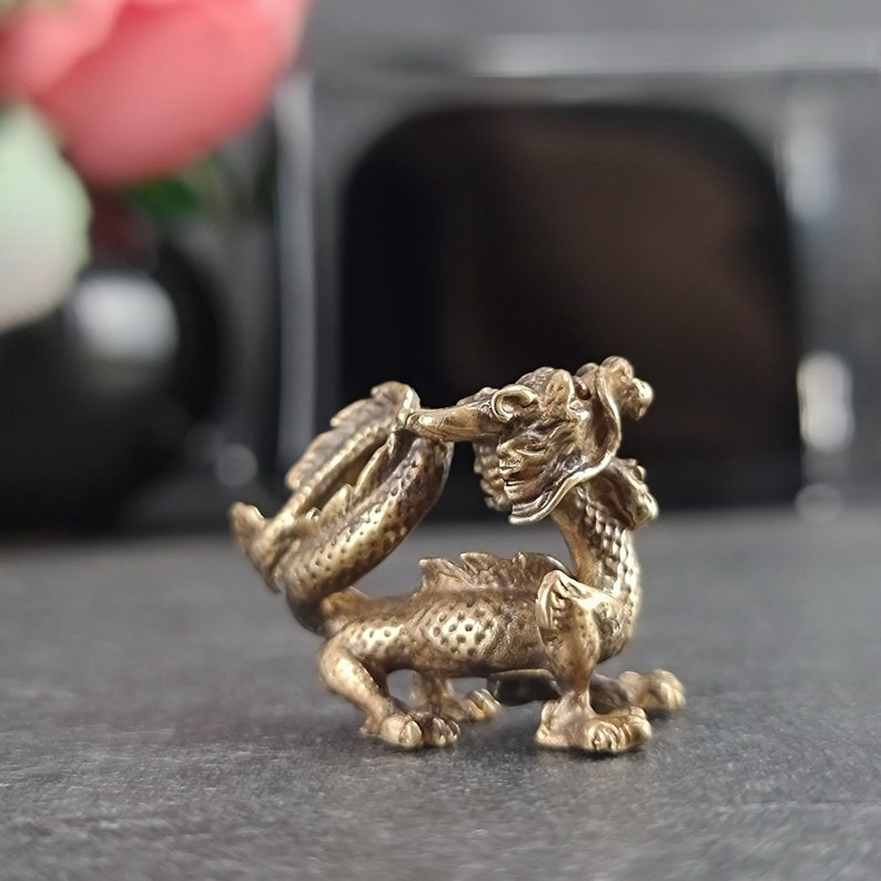 May include: A small, gold-colored brass dragon figurine with intricate details. The dragon is curled up with its tail in its mouth.