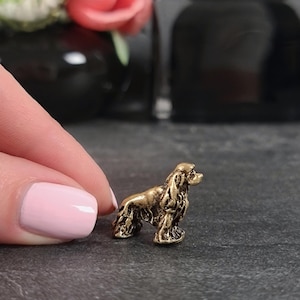 Puede incluir: Pequeña figura de latón de un perro Cocker Spaniel. El perro está representado de pie, con detalles intrincados de su pelaje y rasgos. La figura tiene un cálido acabado dorado antiguo.