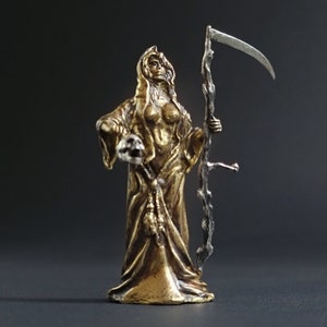 May include: A gold-colored figurine of a female figure in a hooded robe, holding a skull in one hand and a scythe in the other. The figure is standing on a black surface.