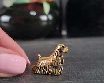 May include: A miniature brass Cocker Spaniel figurine. The dog is shown in a seated pose, with its distinctive long ears and tail clearly visible. The figurine has a polished, golden appearance and is likely intended for decorative purposes.