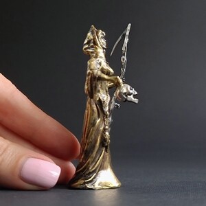 May include: A gold-colored figurine of a woman in a hooded robe holding a scythe and a skull. The figurine is about 6 inches tall and is made of metal.