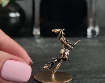 Puede incluir: Una pequeña figura de bronce de una bruja montando una escoba. La figura está sobre una base redonda de oro.