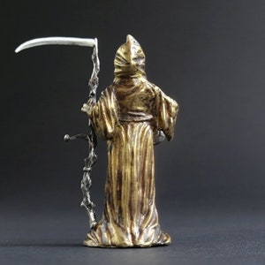 May include: A gold-colored figurine of the Grim Reaper holding a silver scythe. The figurine is standing on a black surface.