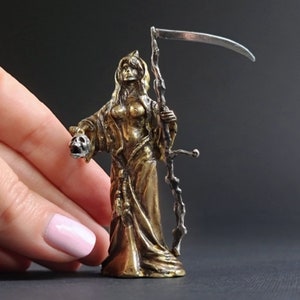 May include: A gold-toned figurine of a woman holding a skull in one hand and a scythe in the other. The woman is wearing a long robe and has a hood over her head.