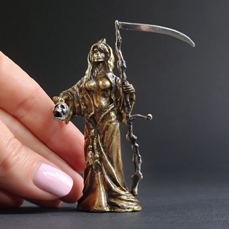May include: A gold-toned figurine of a woman holding a skull in one hand and a scythe in the other. The woman is wearing a long robe and has a hood over her head.