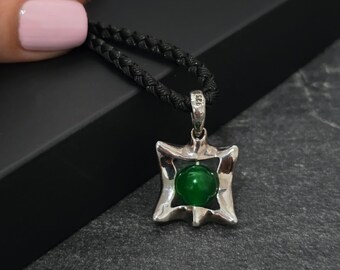 May include: A silver pendant with a green gemstone center, suspended from a black braided cord. The pendant has a unique, stylized square shape with a polished finish. The cord is knotted and appears durable.