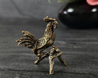May include: A detailed bronze-colored rooster figurine perched on a small, rustic stand. The rooster has intricate feather detailing and a prominent comb. The stand has a three-legged design, adding to the figurine's charm.