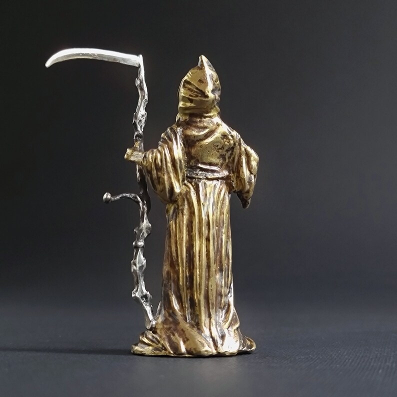 May include: A gold-colored figurine of Death, wearing a hooded robe and holding a silver scythe. The figurine is standing on a black background.