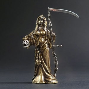 May include: A gold-colored metal figurine of a female figure holding a skull in one hand and a scythe in the other. The figure is wearing a long robe and has long hair. The figurine is standing on a black surface.