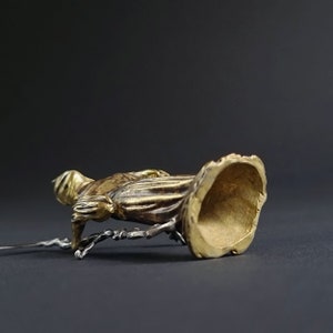 May include: A gold-colored metal figurine of a person in a flowing robe. The figurine is standing on a small, round base. The base is hollow and has a smooth, polished finish.