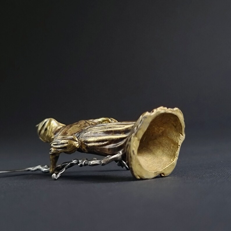 May include: A gold-colored metal figurine of a person in a flowing robe. The figurine is standing on a small, round base. The base is hollow and has a smooth, polished finish.