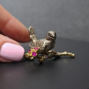 May include: A silver bird figurine perched on a branch with ruby red gemstones. The bird is detailed with a textured finish and has a slightly open beak.