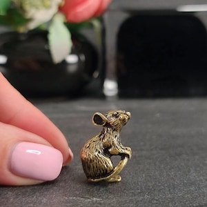 Peut inclure: Une petite figurine de souris en métal de couleur dorée. La souris est assise sur ses pattes arrière, sa queue enroulée autour de son corps.