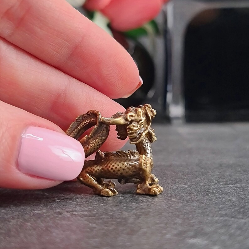 May include: A small, gold-colored dragon figurine with intricate details. The dragon is curled up with its tail wrapped around its body. The figurine is made of metal and has a textured surface.