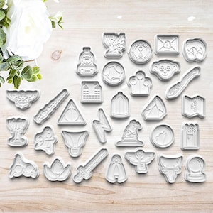 May include: A set of 25 white plastic cookie cutters featuring Harry Potter themed designs, including a Hogwarts Express train, a golden snitch, a Sorting Hat, a lightning bolt, a house crest, a broom, and more.