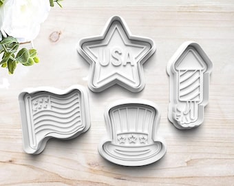 May include: Four white plastic cookie cutters in patriotic shapes: a star with "USA" text, a waving American flag, a top hat with stars, and a firework. These cutters are ideal for creating themed cookies for Independence Day or other patriotic events.