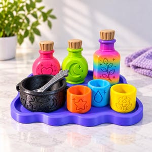 May include: A purple tray holds a black cauldron with a spoon, three small bottles with tan corks, and three colorful cups. The bottles feature heart, moon, and leaf designs. The cups display star, water drop, and flower icons in orange, blue, and yellow.