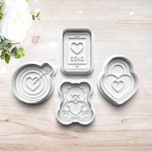 May include: Four white cookie cutters in various shapes: a heart bullseye, a smartphone with a heart and the word "SEND", a teddy bear holding a heart, and a heart-shaped padlock. Perfect for Valentine's Day or other romantic occasions.