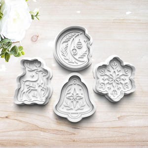 May include: Four white plastic cookie cutters in holiday shapes: a reindeer, a crescent moon with ornaments, a bell, and a snowflake. The cutters are arranged on a light wood surface, perfect for festive baking.