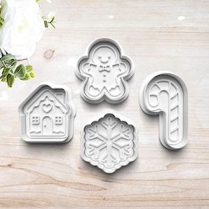 May include: Four white plastic cookie cutters in holiday shapes: a gingerbread man, a house, a snowflake, and a candy cane. The cutters are arranged on a light wood-grain surface, perfect for festive baking.