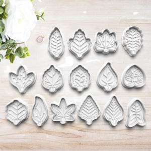 May include: A set of 15 white plastic leaf-shaped cookie cutters. The cutters are arranged in two rows on a light brown wooden surface.