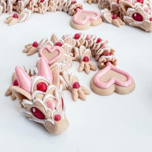 May include: Three pink and white ceramic dragon figurines with heart-shaped details. The dragons are decorated with white icing and pink hearts.