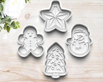 May include: Set of four white plastic Christmas cookie cutters. The cutters are shaped like a gingerbread man, a snowman, a star, and a Christmas tree.